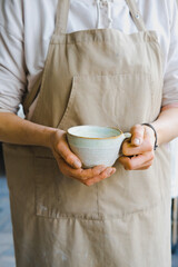 person in the apron holding handmade clay and turquoise cup of tea