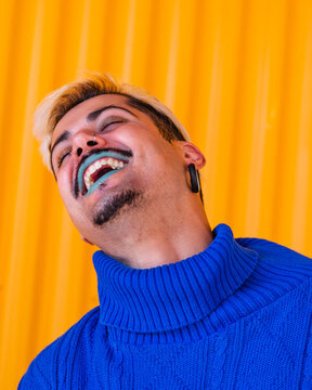Androgynous cheerful male with blue lips and in stylish wear standing on background of yellow wall in city with eyes closed