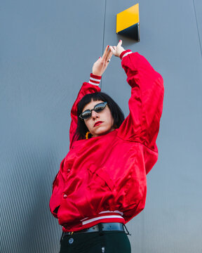 Low angle of female millennial wearing vivid red jacket and stylish sunglasses standing with raised arms on street and looking at camera
