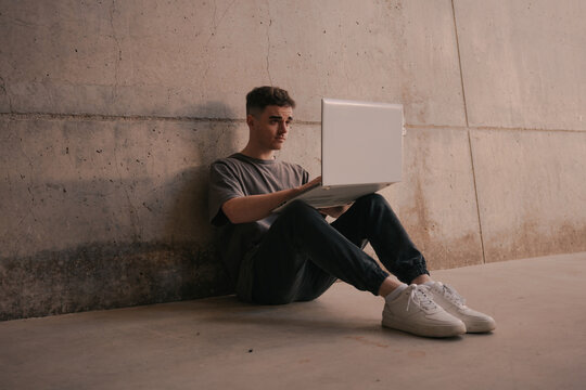 Full length of serious young self employed guy in trendy outfit using laptop while sitting on ground leaning on concrete wall during remote job