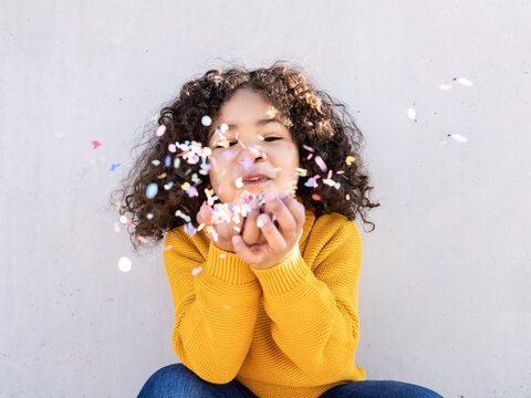 Naklejki Cheerful ethnic child with curly hair blowing colorful paper confetti at camera on sunny day against gray wall