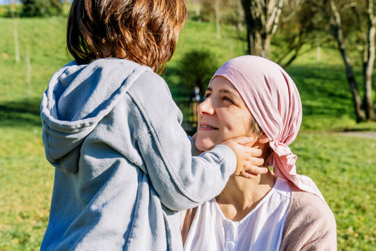 Unrecognizable daughter drawing smile with fingers on mother's face with cancer wearing pink head scarf standing on green park looking at each other