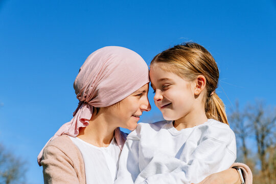 Happy mother with cancer wearing pink head scarf embracing little daughter on green park looking at each other