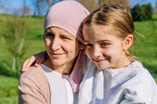 Happy mother with cancer wearing pink head scarf embracing little daughter on green park looking away