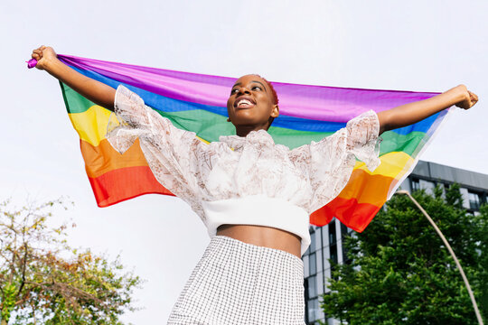 From below of delighted African American lesbian female standing with rainbow LGBT flag on city street and looking away
