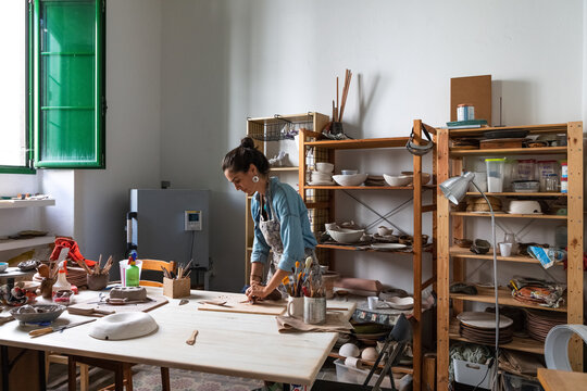 Focused adult lady in casual clothes and apron sculpting clay piece and smiling during pottery class in workshop