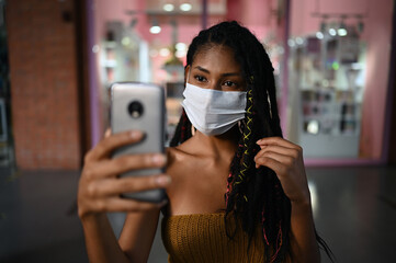 Portrait of attractive young afro latin woman wearing a facemask takes selfie with smartphone in a commercial mall, Colombia