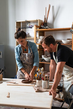 Positive middle aged female and young male in casual clothes and aprons smiling while creating clay plate during pottery class in workshop