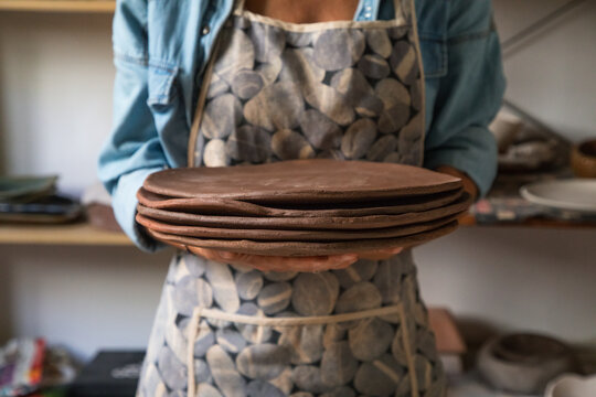 Crop unrecognizable female artisan in casual outfit and apron holding stack of flat clay plates while working in art studio