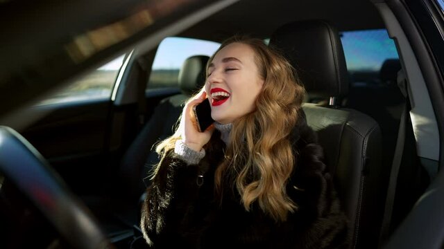 Cheerful happy beautiful woman talking on the phone and laughing sitting in car on driver's seat. Side view of joyful Caucasian gorgeous brunette lady having fun in vehicle. Slow motion.