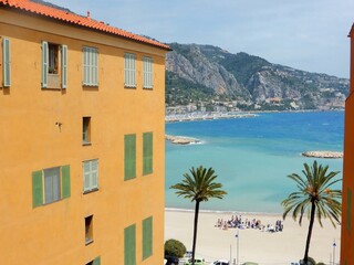 Façade d'immeuble jaune dans la ville de Menton, avec vue sur la plage des Sablettes et l’eau bleu turquoise de la mer Méditerranée, sur la côte d'azur, dans les Alpes-Maritimes, en Provence (France)