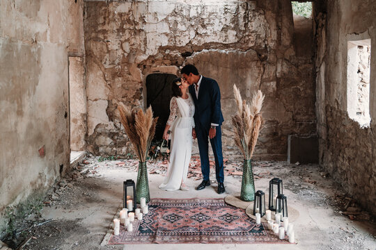 Side view full body loving young ethnic newlywed couple wearing chic wedding clothes embracing and kissing gently in obsolete remaining building