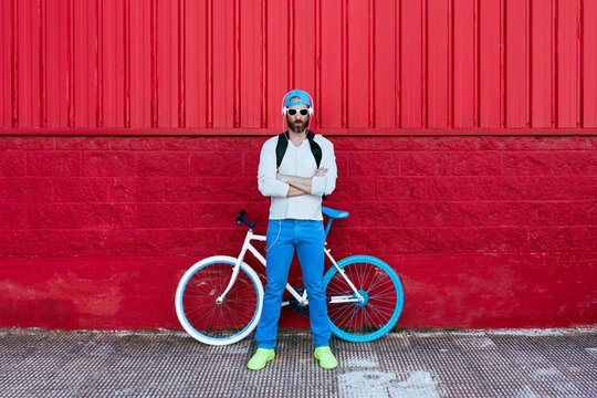 Serious bearded hipster man standing with arms crossed with modern bike on colorful red wall on the street