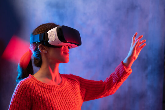 Unrecognizable young female in casual wear and VR headset getting new experience and touching virtual object in room with colorful projector illumination