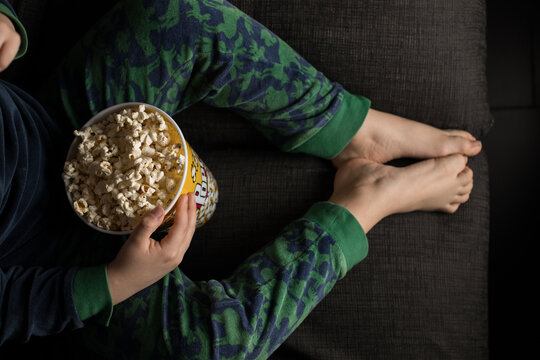From above cropped unrecognizable kid sitting comfortable on sofa with popcorn bucket while eating and watching TV