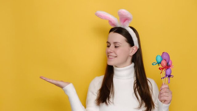 Beautiful young girl holding little colorful eggs is pointing with raises palm and looking on empty copy space, wears pink bunny ears and jumper, isolated on yellow studio background. Easter concept