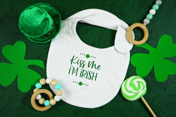 Happy St Patrick's Day baby wear white bib, styled with leprechaun hat, shamrocks, on a textured...