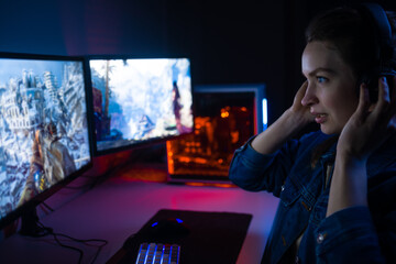 A girl with headphones plays a video game on a computer on large monitors. Gamer with mouse and keyboard. Online games with friends,win.Fun entertainment.Teenagers play adventure games.Neon lighting