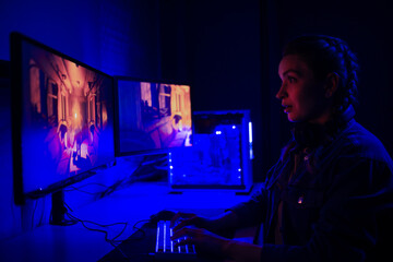 A girl with headphones plays a video game on a computer on large monitors. Gamer with mouse and keyboard. Online games with friends, win.e-sports, streams.Teenagers play adventure games.Neon lighting