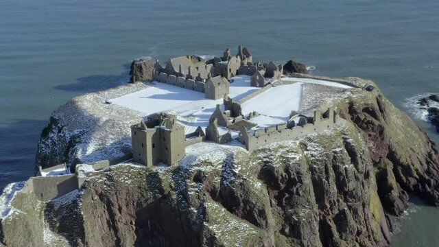 Aerial view of Dunnottar Castle ruins surrounded by snow on a sunny winter day, nr Stonehaven, Aberdeenshire, Scotland. R to L rotating CU of the south and west sides of the castle whilst zooming out.