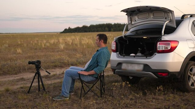 A man traveler in car stopped at camping in evening and takes pictures with a camera next to car. Freelance photographer working outdoors, beautiful moments for photos and videos. Driver is resting