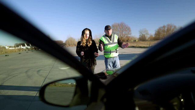 Wide shot of man in green vest explaining driving to tired young woman on sunny day outdoors. Exhausted beautiful Caucasian lady studying in driving school. Professional auto instructor teaching.
