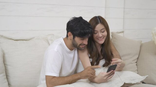 A couple family. Happy middle east young man and Asian woman. People using a smartphone on social media internet and sitting on safa at home. Lifestyle in technology device. Multi ethnic