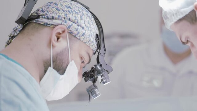 Doctor and an assistant in the operating room for surgical venous vascular surgery clinic. Action. Side view of a surgeon with a binocular loupe.