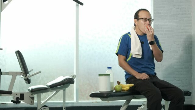Young Asian man in sportswear sitting on bench esting red apple feel good after weight training in Gym. sport, bodybuilding, fitness and people concept