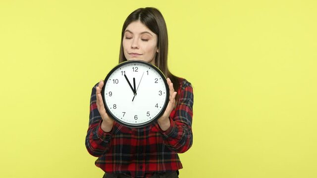 Happy satisfied brunette woman in checkered shirt showing big wall clock looking at camera with smile, satisfied with completed on time work. Indoor studio shot isolated on yellow background