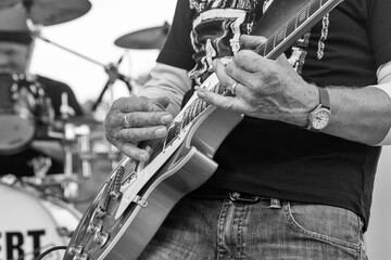 Black and white close up of guitar playing with drum set in the background