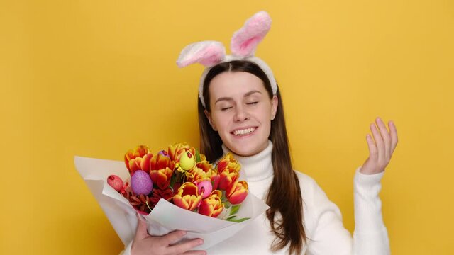 Cheerful woman in bunny fluffy ears hold bouquet of spring flowers decorated with Easter colored eggs enjoying good smell or pleasant fragrance, feel calm breathe fresh air, isolated on yellow wall
