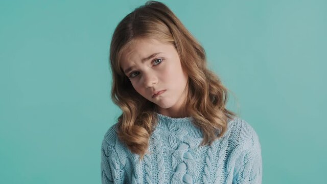 Upset blond teenage girl feeling dejected sadly posing on camera isolated on blue background. Guilty expression