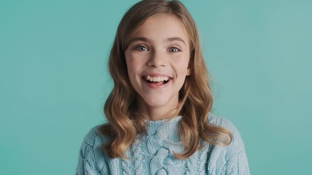 Pretty excited blond teenage girl looking surprised on camera and smiling over blue background