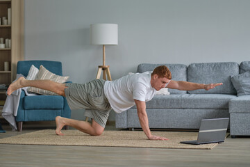 redhead man in white t-shirt works out in the living room. Man doing sports on a mat watching...