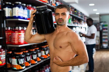 Bare-chested athletic man keeping with cans of sports nutrition in store, demonstrating muscles.