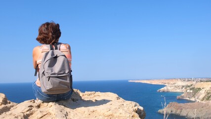 Fototapeta na wymiar Young happy woman with a backpack sits on a rock and looks at the valley below. Tourists on the rocky coast of the Black Sea. The concept of freedom and ease