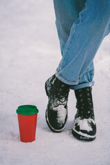 Fototapeta premium Red paper cup stands on the snow next to feet in snowy boots. Mock up, copy space. Walk in the snow