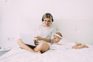 In the bedroom, a young beautiful blonde is working on a white bed in headphones with a laptop, next to her is a little 6-month-old daughter.
