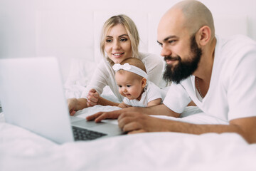 Fototapeta premium A beautiful Caucasian family mom, daughter and bearded dad lie on the bed in the bedroom in the morning and watch cartoons on a laptop. Parents and children.