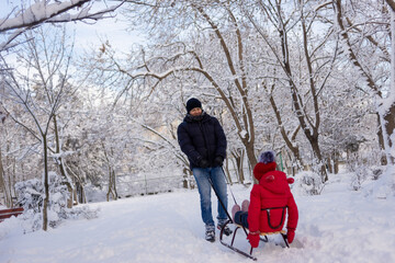 Dad is taking his daughter, a schoolgirl in a red jacket, through the winter park on a sleigh. Rest and time in the fresh air.