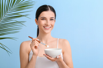 Beautiful young woman with bowl of facial cream, brush and tropical leaf on color background