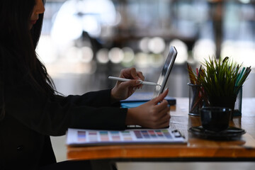 Side view of young female designer holding stylus pen drawing on digital tablet while sitting in creative office room.
