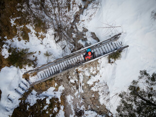 aerial drone shot of hiker with backpack, orange hat and orange pants standing next to waterfall surrounded by icicles and snow in winter in austria