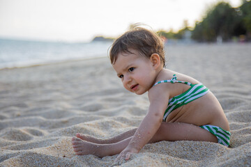 Fototapeta na wymiar adorable toddler in a swimsuit sits on a sandy beach in the sunshine.