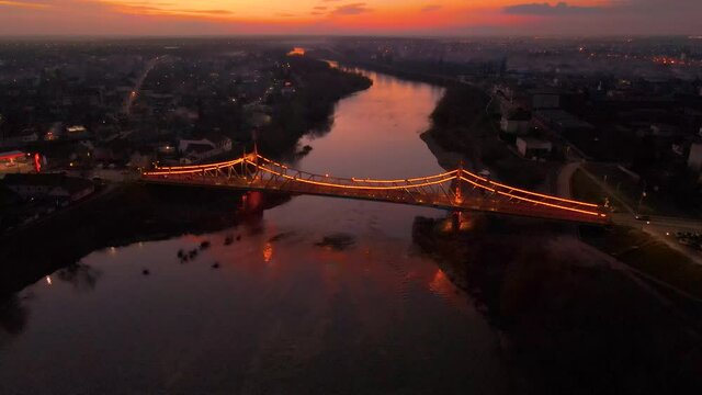 Drone footage of a brightly lighted spanning bridge over a river at sunset. Drone flying backwards and picking up altitude keeping the bridge in focus. Illuminated bridge at sunset filmed from a drone