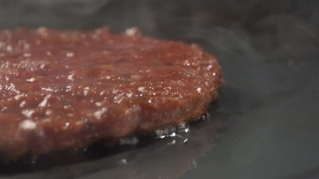 Steam comes from hot skillet surface and bubbling oil frying patty of meat. Hamburger cooking in kitchen with warm juices from fat and oil sizzling. Close up view artificial or fake meat cooking.