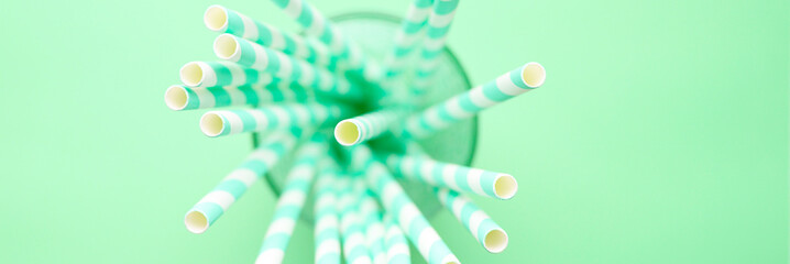 pile of paper striped white and green drinking straws for party in clear glass cup on green background. banner
