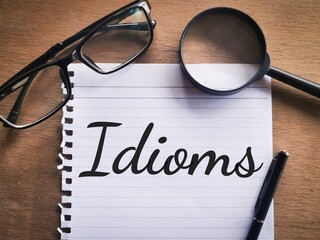 Phrase IDIOMS written on a piece of paper with a pen,eye glasses and magnifying glass. Motivational...