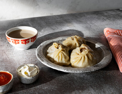 Buuza in a ceramic plate next to a red napkin with a pattern. Seasoning sauce of tomatoes with garlic, sour cream and a bowl of green tea. National, traditional Buryat-Mongolian dish.
